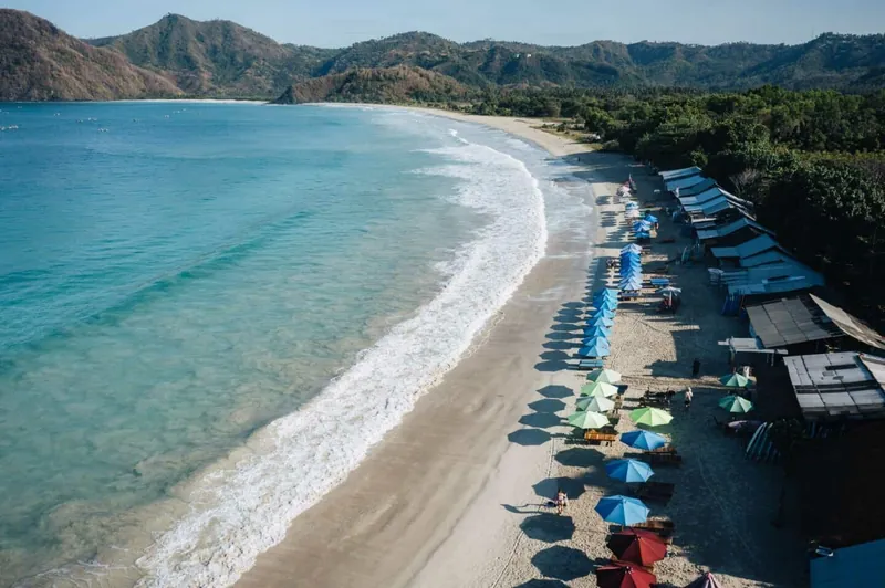 Aerial view of Selong Belanak Beach in Lombok with turquoise water, gentle waves and a line of colorful umbrellas and beach huts.