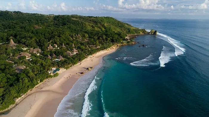 Aerial view of Nihi Sumba resort hidden in lush hills above a long empty beach on Sumba, Indonesia.