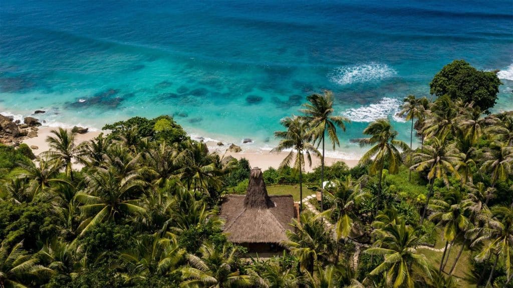 Traditional villa surrounded by palm trees on a secluded beach with turquoise ocean in Sumba, Indonesia.